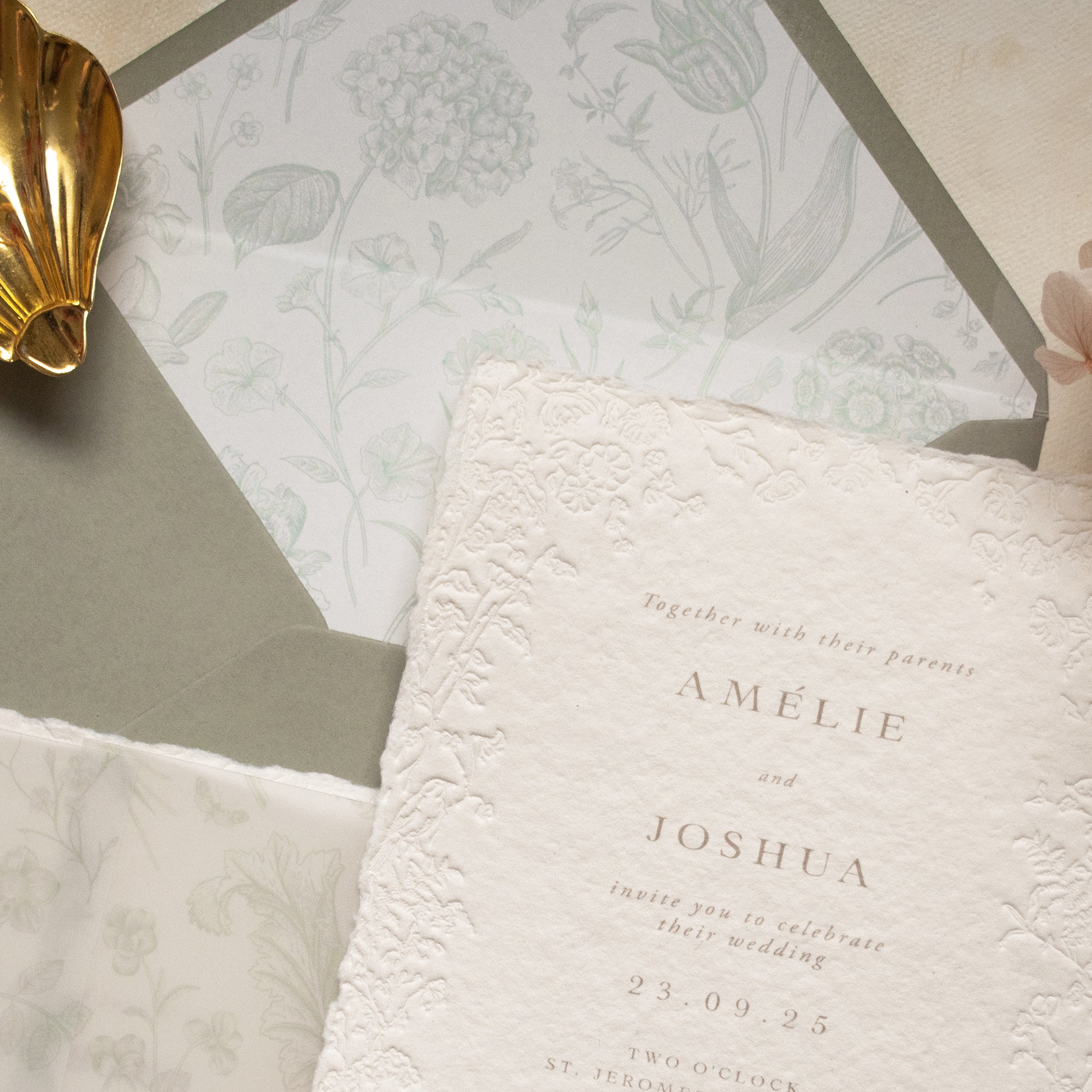A close-up of a white textured paper invitation card with blind letterpress floral borders, layered over a dusty sage floral envelope