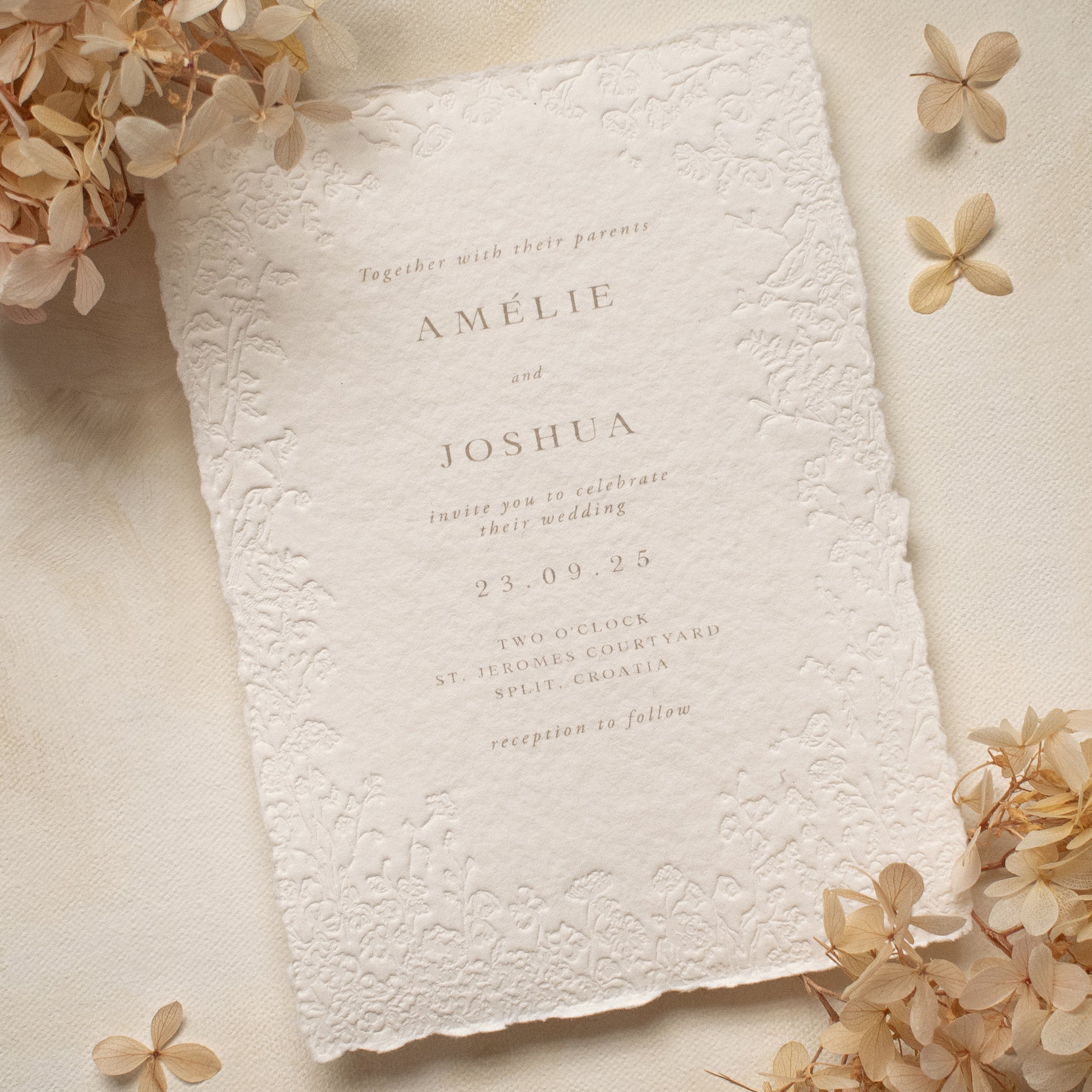 A close-up of a white textured paper invitation card with blind letterpress floral borders, styled with beige dried flowers